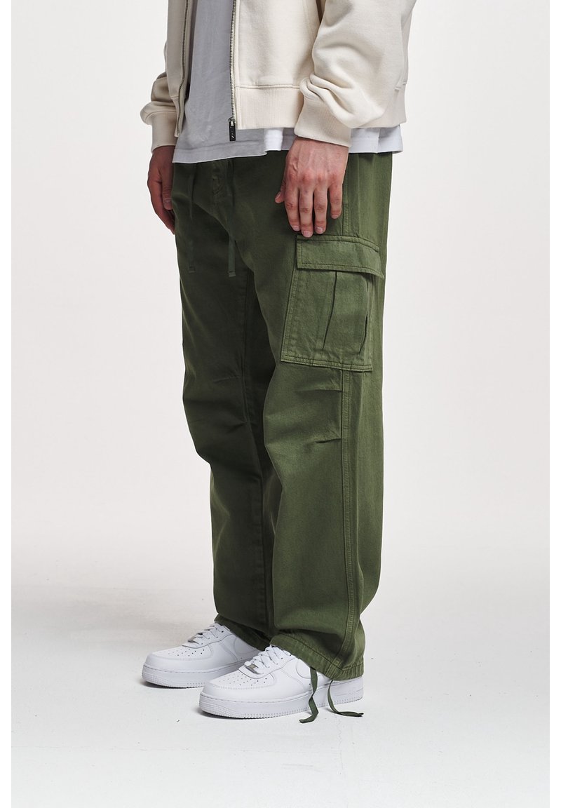 Personne portant un pantalon cargo vert olive avec de grandes poches latérales, des baskets blanches et une veste zippée beige clair.