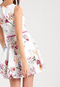 La robe à imprimé floral présente une base blanche, un design sans manches et une ceinture rose à la taille. La jupe s’évasent avec des motifs floraux vibrants.
