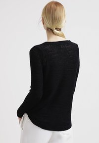 Schwarzer Strickpullover mit langen Ärmeln, einem tief ausgeschnittenen Rundhalsausschnitt und einem etwas längeren Saum auf der Rückseite. Texturiertes Material mit lockerem Gewebe.