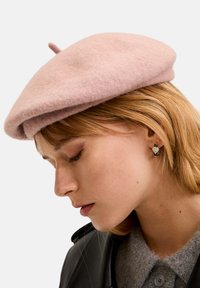 Hellrosa Wollberet mit runder Form und einem kleinen oberen Tab. Begleitet von einem grauen Pullover mit Kragen und Ohrsteckern.