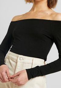 Frau trägt ein schwarzes, schulterfreies Langarmoberteil, das in eine hochgeschnittene beige Hose mit Knopfdetails gesteckt ist.