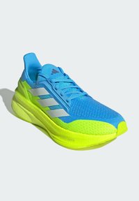 Zapatilla de running Adidas en azul y amarillo neón con franjas blancas, suela ligera boost y cordones sobre un fondo claro.