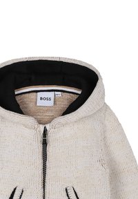 Felpa in maglia beige con interno nero, chiusura a zip e dettagli neri a contrasto sulle tasche. Presenta un tessuto testurizzato e morbido.