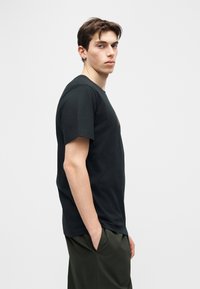 Νεαρός άνδρας με σκουλαρίκια, φοράει μαύρο κοντομάνικο μπλουζάκι και σκούρο πράσινο παντελόνι, στέκεται πλαγίως με τα χέρια στις τσέπες.