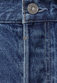 Denim-kangas tummansinisellä sävyllä, jossa näkyvät kulutukset, kuparinvärinen metallinappi, jossa lukee "ALLSAINTS", sekä pieni hopeavälinen etiketti vyötäröllä.