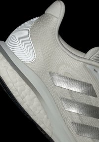 Hellgraue Sportasche mit einer strukturierten Oberseite, metallisch silbernen Streifen, Gummisohle mit Wellenmustern und einem gepolsterten Kragen.