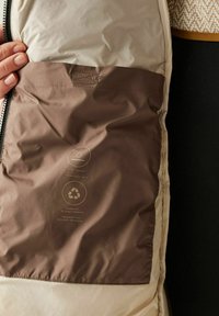 Mano sostiene abierta una chaqueta beige que revela un bolsillo interior marrón etiquetado como repelente al agua y hecho con materiales reciclados en inglés y alemán.