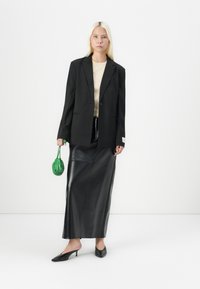 Czarny oversize blazer na beżowej bluzce, zestawiony z elegancką czarną skórzaną spódnicą. Modelka trzyma małą zieloną torebkę. Czarne czółenka na niskim obcasie dopełniają stylizację.