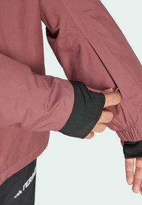 Letvægts, mauve jakke med elastiske manchetter; har et sort tommelhul og en lynlåslomme på ærmet. Glat, mat overflade.
