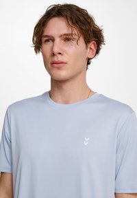 Ljusblå kortärmad t-shirt tillverkad av mjukt tyg, med en liten vit chevronlogotyp på vänster bröstområde.