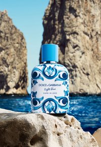 Perfume bottle with blue floral design labeled Dolce & Gabbana Light Blue Capri in Love placed on rock by sea with large cliffs in background.