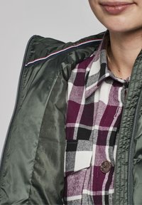 Zelená bunda na zip s lesklou tkaninou, s proužkovaným detailem na límci, přes fialovou, černobílou kostkovanou košili se zapínáním na knoflíky.