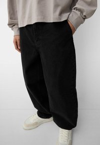 Black, loose-fit denim pants with side pockets and a denim texture, paired with white and beige athletic sneakers.