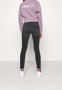 Persona con jeans skinny negros, zapatillas blancas y un sudadera corta en color malva con el texto "NIKE AIR" en la parte de atrás, de pie contra una pared blanca.