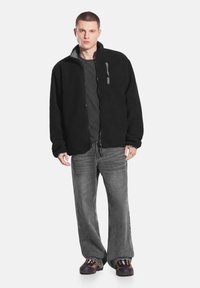 Czarna kurtka polarowa z frontowym zamkiem błyskawicznym i szarymi akcentami, zestawiona z luźnymi szarymi dżinsami oraz czarnymi butami z wzorzystymi detalami.