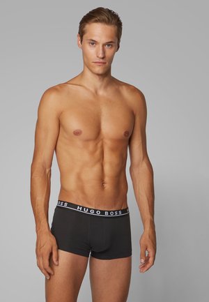 Schwarze Boxershorts mit einem weißen elastischen Bund, auf dem "HUGO BOSS" steht. Das Modell steht mit einem neutralen Ausdruck vor einem grauen Hintergrund.