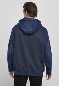 Felpa con cappuccio di colore blu navy, con una texture liscia, caratterizzata da un ampio cappuccio, polsini a costine e un design del pannello laterale. Posizionata su uno sfondo neutro.