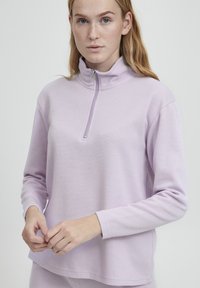Světle levandulový pulovr vyrobený z měkké tkaniny, s polo-zip límcem a dlouhými rukávy v pohodlném střihu. Minimalistické detaily šití.