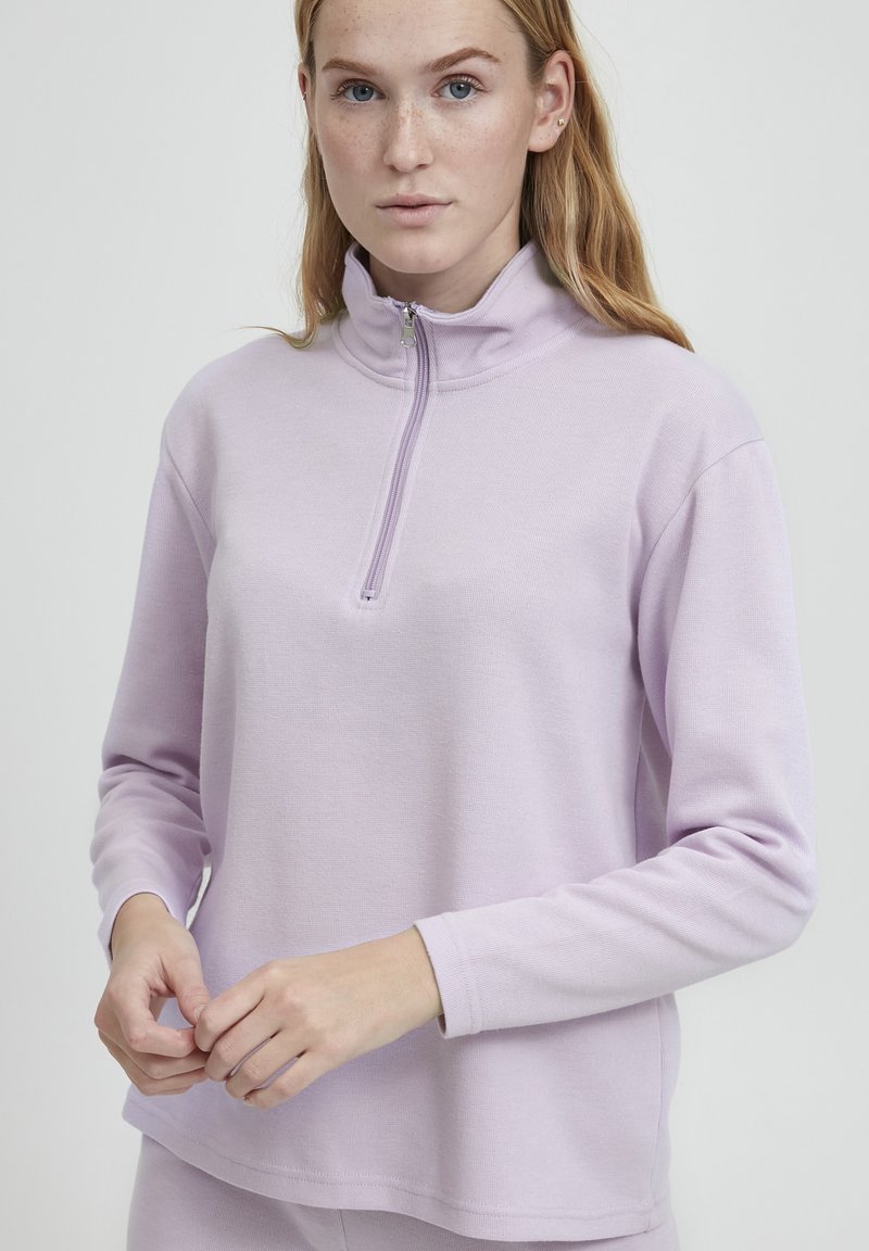 Světle levandulový pulovr vyrobený z měkké tkaniny, s polo-zip límcem a dlouhými rukávy v pohodlném střihu. Minimalistické detaily šití.
