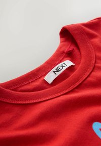 Sarkana kokvilnas T-krekls ar apaļu kakla izgriezumu un nelielu zilu grafisko dizainu. Etiķete ar uzrakstu "NEXT" uz baltas auduma etiķetes. Gluda tekstūra.