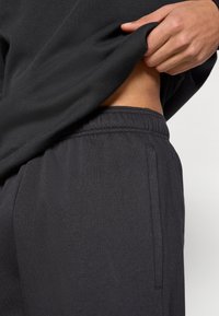 Μαύρο βαμβακερό φούτερ που καλύπτει μέρος σκούρων παντελονιών jogger. Τα joggers διαθέτουν ελαστική μέση και πλαϊνή τσέπη, με λεία υφή.