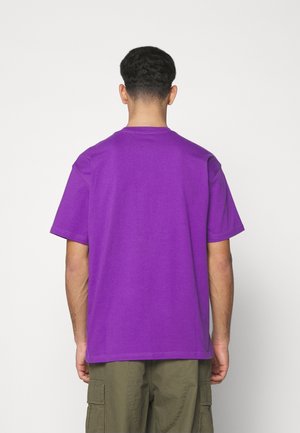 Mann mit kurzen lockigen Haaren von hinten gesehen, trägt ein schlichtes lila T-Shirt mit kurzen Ärmeln und olivgrüne Cargohosen.