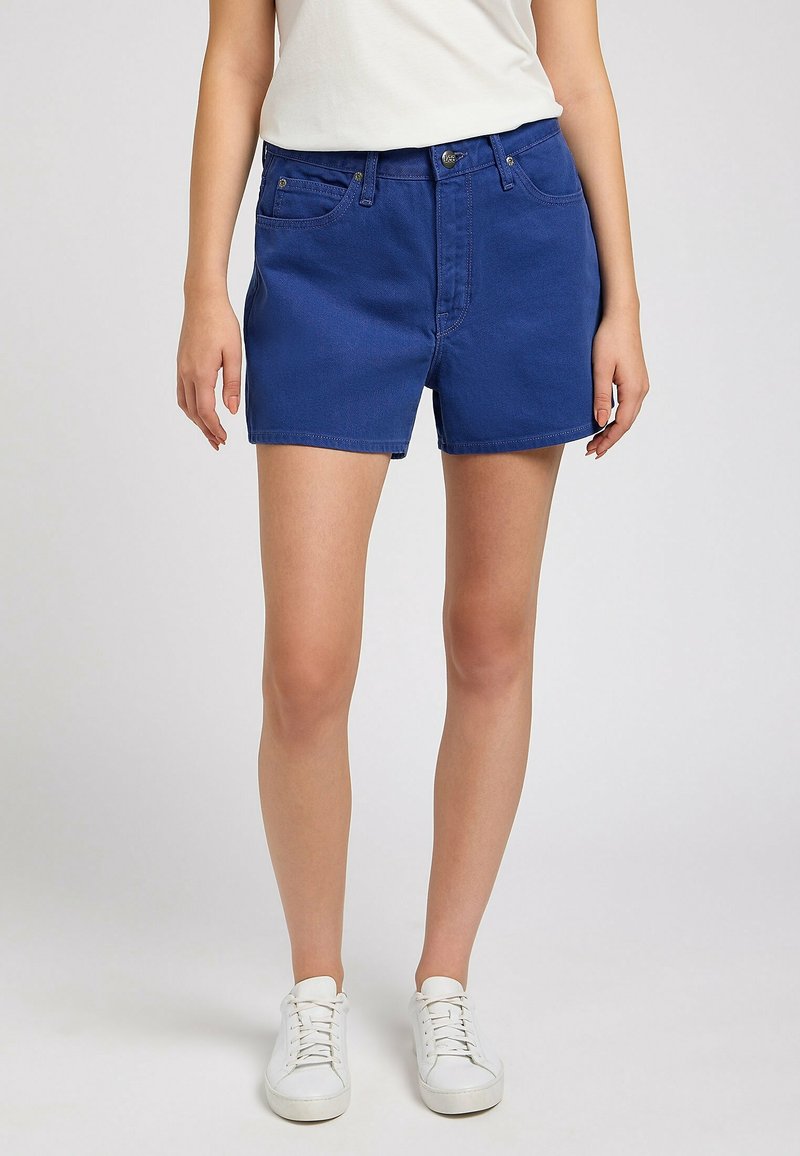 Femme portant un short en denim bleu et des baskets blanches, debout devant un fond clair uni.