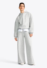 Felpa grigia oversize con un ampio cappuccio e spalle scese, abbinata a pantaloni larghi coordinati. Il tessuto ha una consistenza morbida con una leggera lucentezza.
