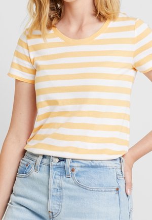 Kvinde iført en gul og hvid vandretstribet T-shirt med korte ærmer stoppet ned i lyseblå højttaljede denimjeans.