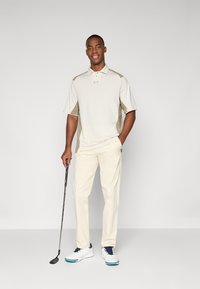 Polo beige avec des accents aux épaules, pantalon crème et chaussures de golf blanches avec des lacets bleus. Tient un club de golf dans une main.
