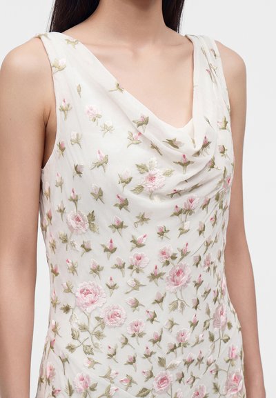 Robe blanche sans manches avec broderie florale rose et verte, avec un décolleté drapé portée par une personne aux longs cheveux foncés.