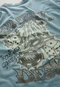 Blauer Stoff mit dem Text "Grateful Dead", zwei Würfel mit Totenkopfgesichtern, knöcherne Handknochen und zwei kreisförmige Blitz-Designs.