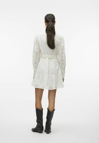 Abito bianco in pizzo con maniche lunghe, vita cinturata e gonna svasata; abbinato a stivali da cowboy neri fino al ginocchio.