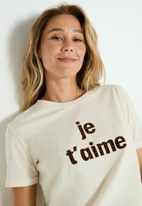 Mujer sonriente con pelo rubio, vestida con una camiseta crema con texto de leopardo "je t'aime", apoyada contra una pared blanca.