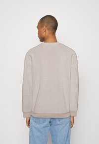 Beige sweater med tekstureret stof, rund halsudskæring og ribbede manchetter, prydet med en kontrasterende brun stribe langs skuldrene og siderne.