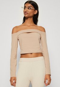 Beige off-shoulder langermet topp med horisontal halsutskjæring; glatt tekstur. Kombinert med kremfargede ribbede bukser. Gjennomsiktige briller brukt.