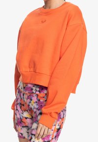 Sudadera cortada de color naranja brillante con mangas anchas y dobladillo acanalado. Combinada con leggins de estampado floral multicolor.
