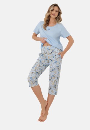 Kobieta stoi boso, ubrana w jasnoniebieski krótki rękaw i spodnie capri w kwiatowy wzór, lekko podnosi koszulkę jedną ręką.