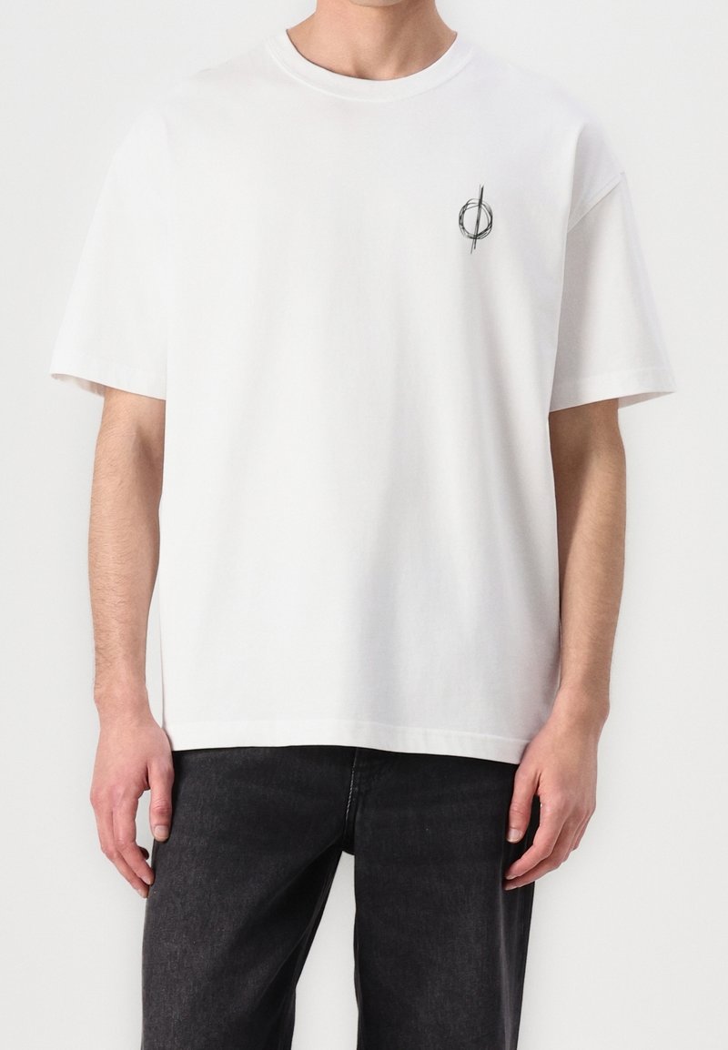 Personne portant un t-shirt blanc ample avec un petit logo circulaire noir sur le côté gauche de la poitrine et un jean foncé, debout devant un fond uni.