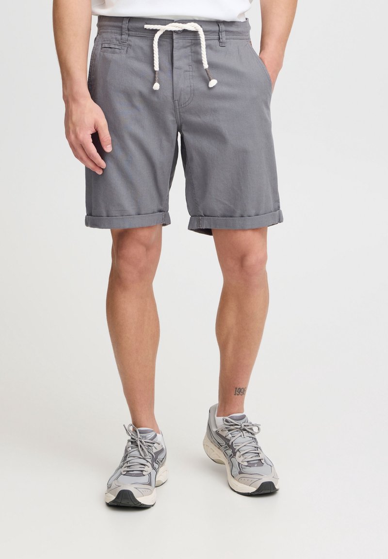 Homme portant un short gris avec ourlet retroussé, ceinturé d'une corde blanche, et des chaussures de course grises, debout les mains dans les poches sur fond blanc.