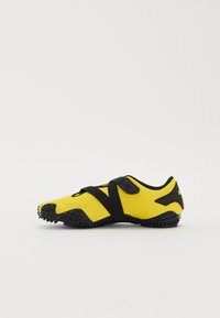 Yellow athletic shoe with black accents, featuring a textured upper, a Velcro strap, and a rubber sole with small protrusions for traction.