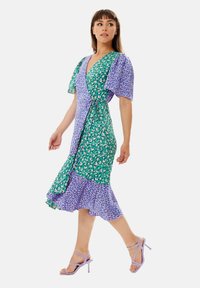 Green and purple patterned wrap dress in lightweight fabric, featuring flutter sleeves, a tie waist, and a flared hem. Paired with lavender heels.