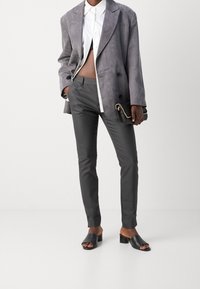 Person trägt grauen Wildlederblazer über weißem, gekürztem Hemd, eng geschnittene graue Hose, schwarze, mules mit Absatz und hält eine schwarze Clutch mit Kettenriemen.