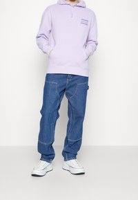 Ljuslila huvtröja med framficka, kombinerad med blå baggy jeans och vita sneakers. Tyget verkar mjukt; jeansen har kontrasterande sömmar.