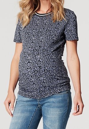 Kobieta w ciąży ubrana w dopasowaną, krótkim rękawem, granatową bluzkę w panterkę i jasnoniebieskie dżinsy, stojąca ze zrelaksowanymi ramionami wzdłuż ciała.