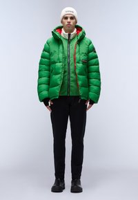 Green puffer jacket with a hood and red accents, layered over a matching zip-up fleece, paired with black pants and black boots.