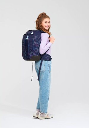 Junge Frau, die lächelt und seitlich steht, trägt einen großen dunkelblauen Rucksack mit geometrischem Muster, ein helllila Oberteil, blaue Jeans und weiße Sneakers.