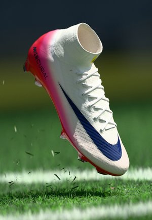 Soccer shoe with a white upper, pink gradient heel, and a blue swoosh. Textured surface and cleats visible against a grassy background.