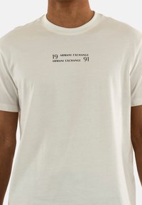 T-shirt blanc à manches courtes en tissu doux, avec un design imprimé portant "19 ARMANI EXCHANGE" et "91 ARMANI EXCHANGE" en noir.