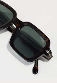 Ochelari de soare cu tortoise brown și lentile verzi închise. Cadru pătrat, accente metalice pe tâmple și branding gravat vizibil pe lentilă.